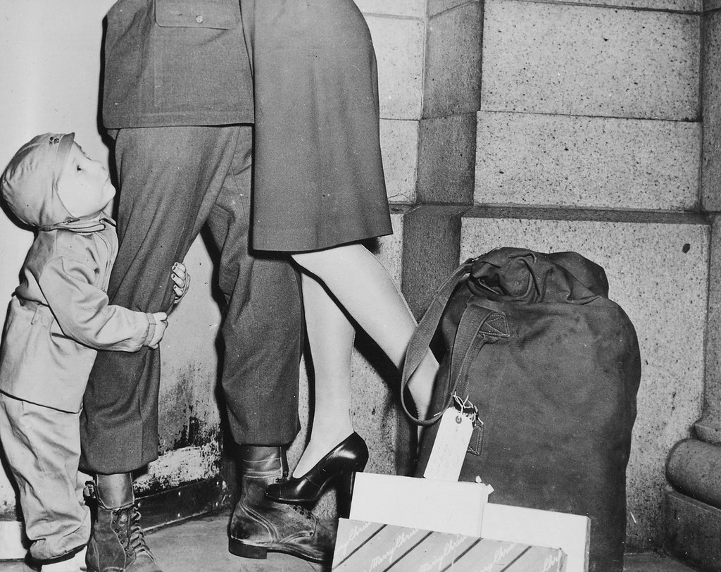 A youngster, clutching his soldier father, gazes upward while the latter lifts his wife from the ground to wish her a Merry Christmas