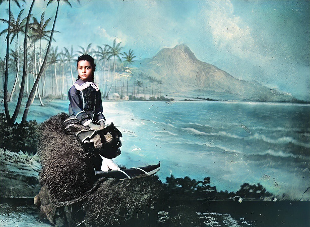 Kaiulani, approximately six years old seated holding hat with backdrop of Diamond Head & palm trees in a photo studio